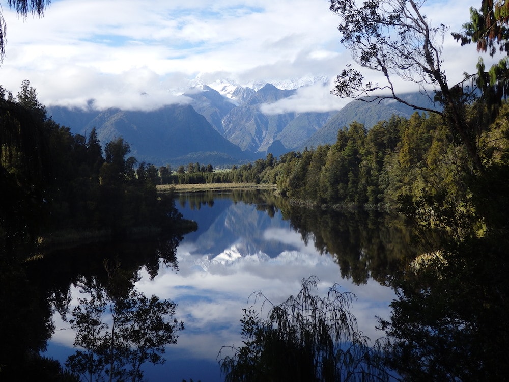 Mountains and clouds reflecting in a lake surrounded by green trees in New Zealand