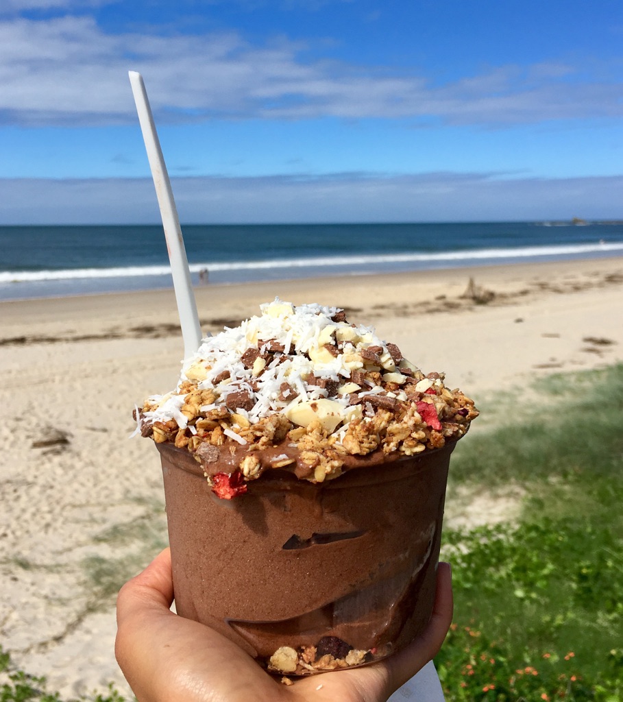 A hand holds an acai bowl with granola and coconut against the backdrop of the beach 