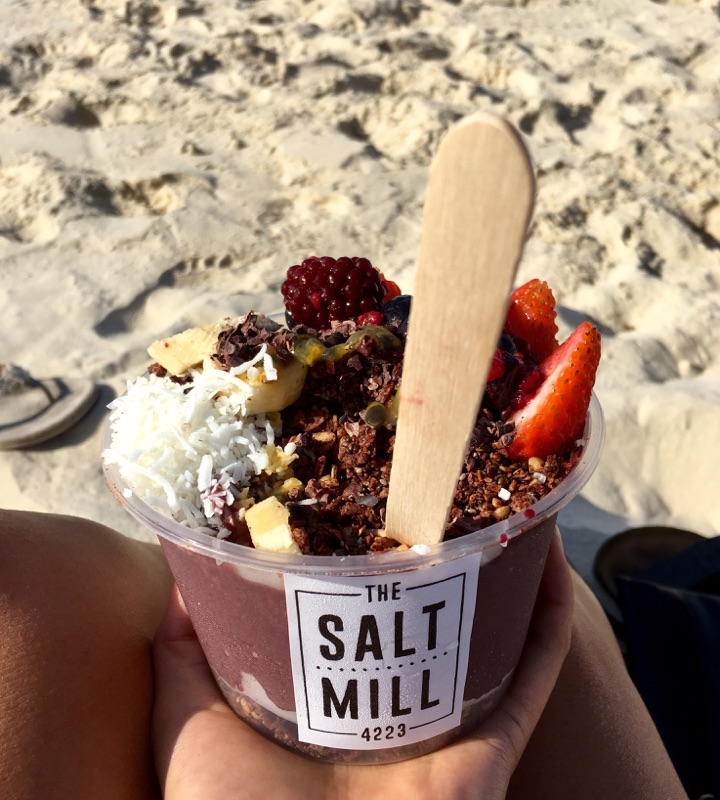 A hand holds an Acai bowl topped with coconut, strawberries and granola on the beach