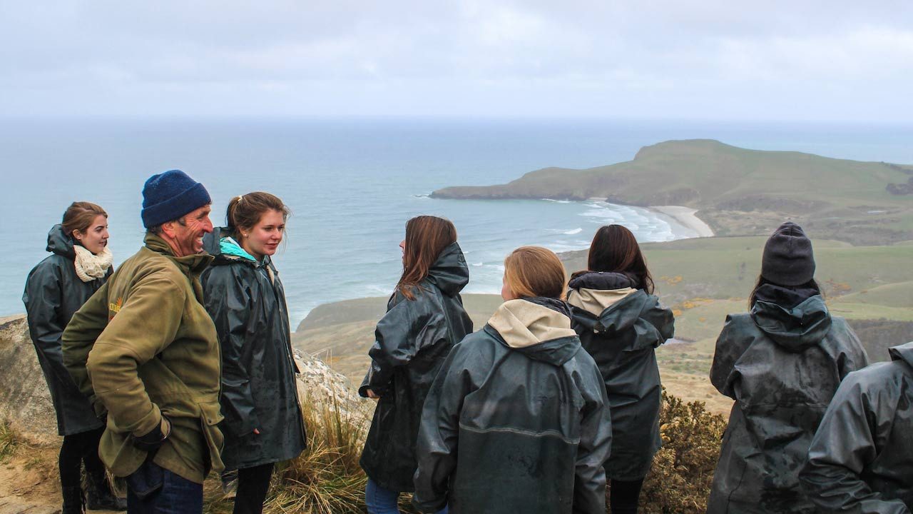 A group of people stand on a mountain top looking out over the ocean near Dunedin, New Zealand