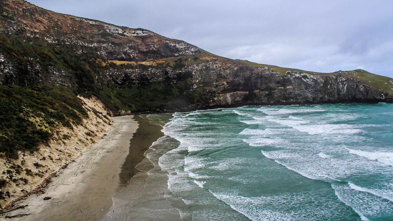 Waves crash to shore beneath surrounding mountains near Dunedin, New Zealand