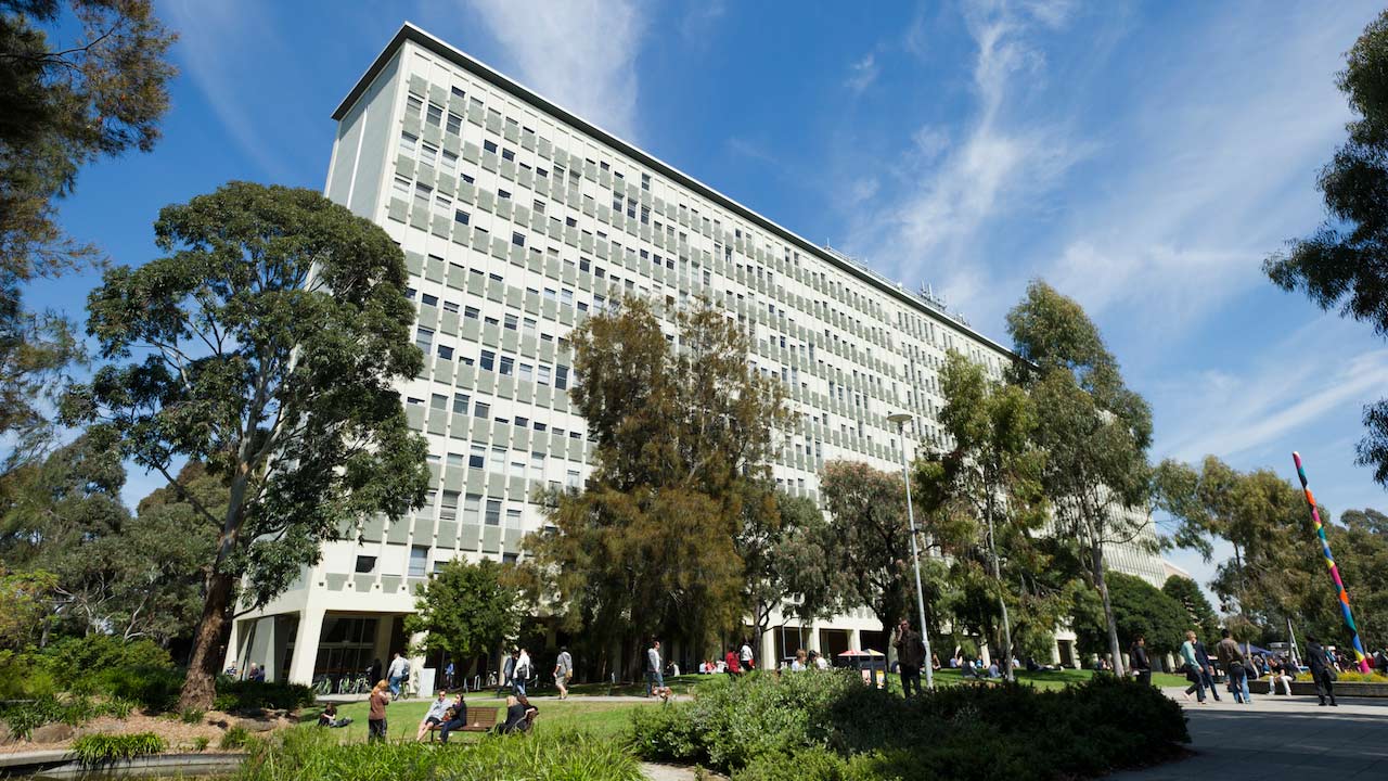 People walk on the pathway and sit on a bench on Monash's campus beneath a large gray building