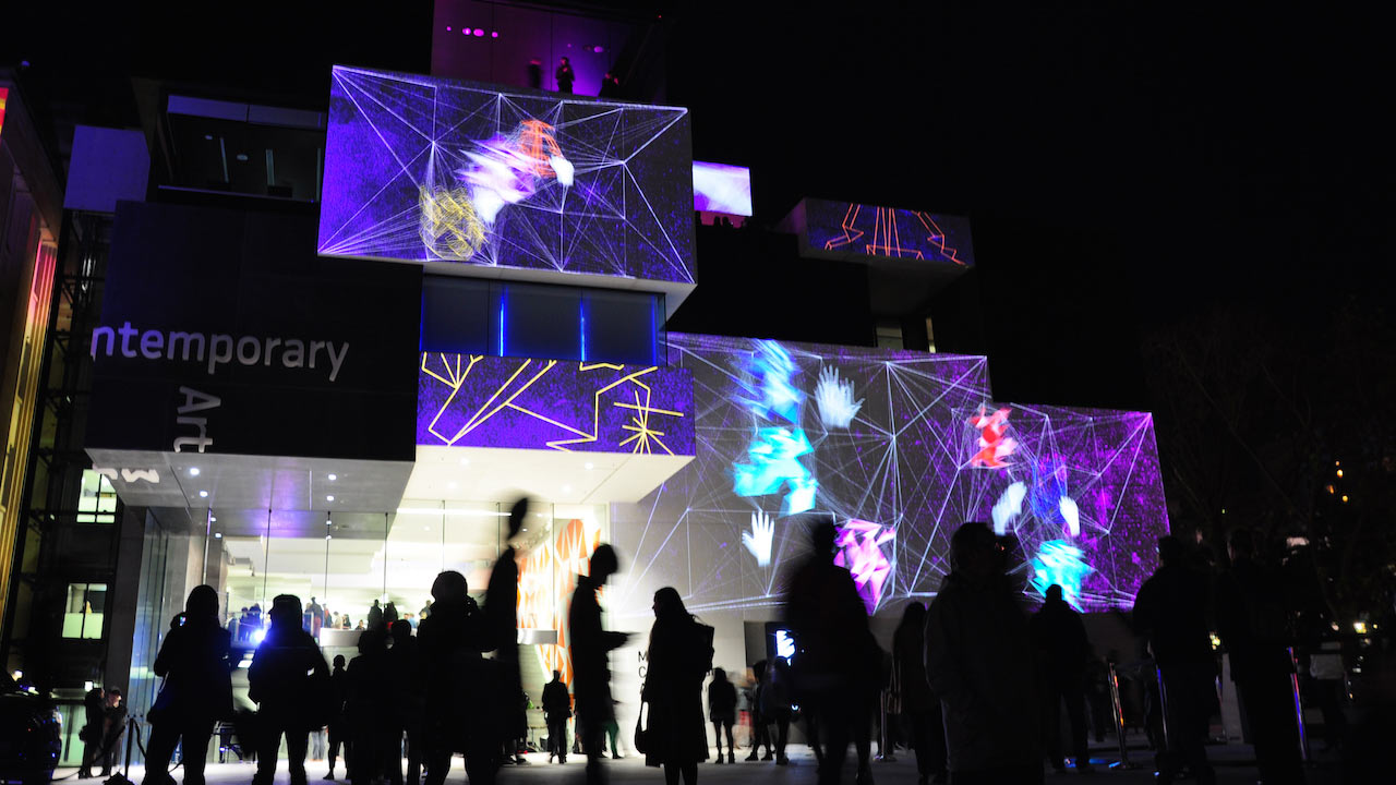 A colourfully illuminated building casts shadows of the crowds of people standing outside in Sydney