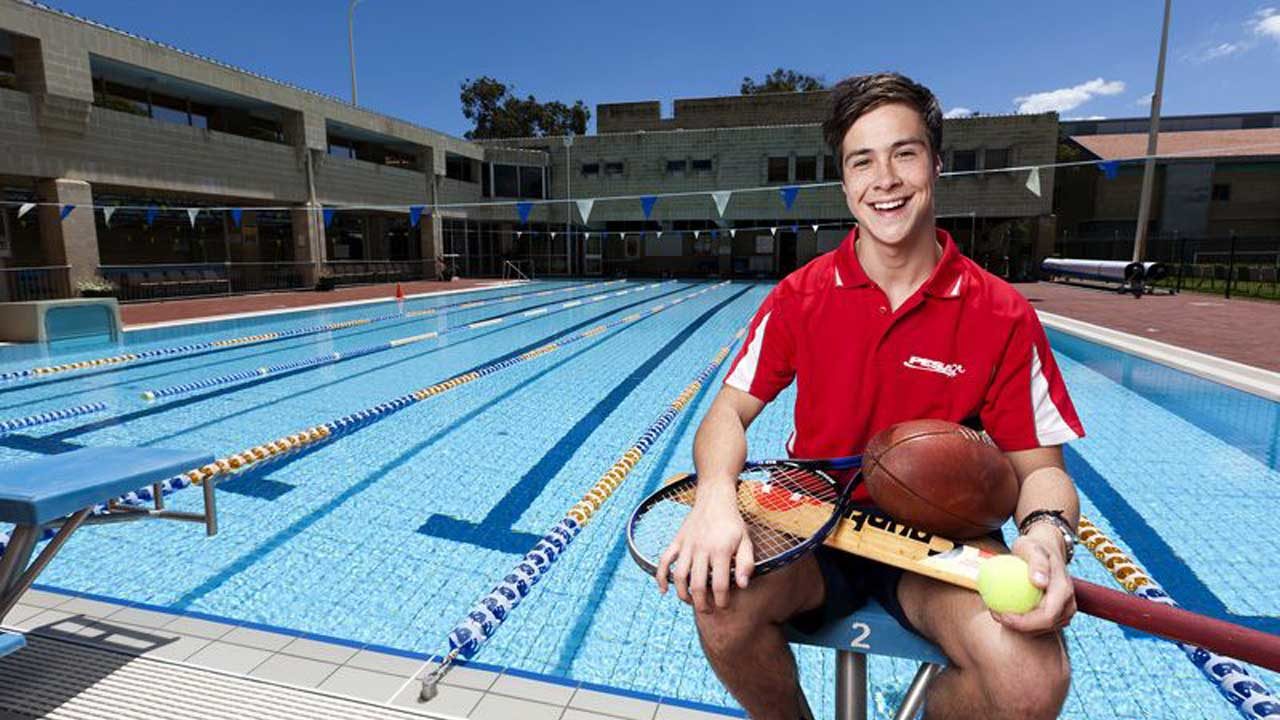 A man sits in front of an outdoor lap pool, smiling, holding an array of athletic gear