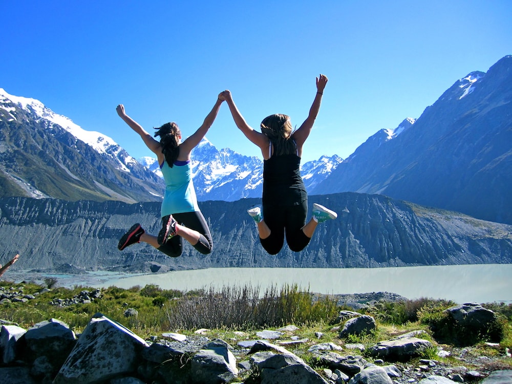 Two friends jumping up in the air with mountains and a lake in the background in New Zealand